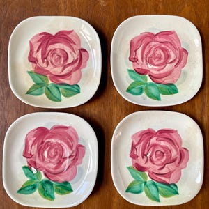 May include: Four square ceramic plates with rounded corners. Each plate features a hand-painted pink rose with green leaves on a cream-colored background. The plates are arranged on a wooden surface.
