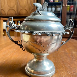 May include: A silver sugar bowl with a bird finial on the lid. The bowl has a footed base and two handles. The bowl is decorated with a floral design.