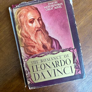 May include: A vintage book titled "The Romance of Leonardo Da Vinci" by Dmitri Merejkowski. The cover features a portrait of Leonardo Da Vinci with a long beard against a maroon background. The book is on a wooden surface.