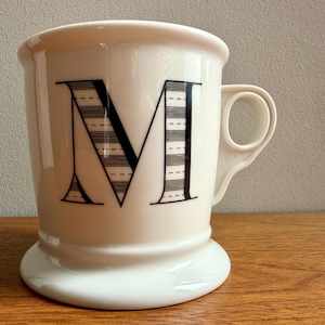 May include: A white ceramic mug with a large black letter "M" on the front. The letter is partially striped with a dotted pattern. The mug has a rounded base and a handle on the side. The mug is sitting on a wooden surface.