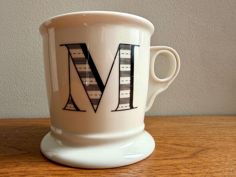 May include: A white ceramic mug with a large black letter "M" on the front. The letter is partially striped with a dotted pattern. The mug has a rounded base and a handle on the side. The mug is sitting on a wooden surface.