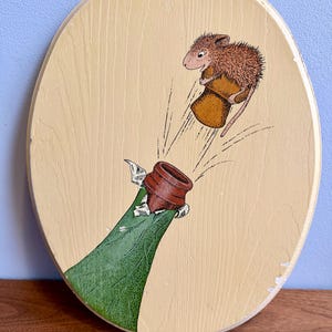 May include: Oval wooden plaque with a whimsical illustration. A brown mouse wearing a brown hat is depicted jumping out of a bottle. The bottle is green with a brown cork. The background is a light beige color.