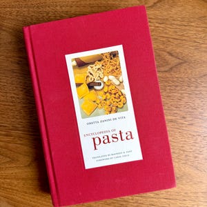 Puede incluir: Un libro rojo de tapa dura titulado "Encyclopedia of Pasta" de Oretta Zanini De Vita. La portada presenta una imagen rectangular de varias formas de pasta. El libro está sobre una superficie de madera.