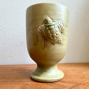 May include: A ceramic tumbler in a muted green hue, featuring a raised relief of a turtle on its surface. The tumbler has a rounded body and a pedestal base, suitable for beverages. The turtle design adds a touch of nature-inspired artistry to the piece.