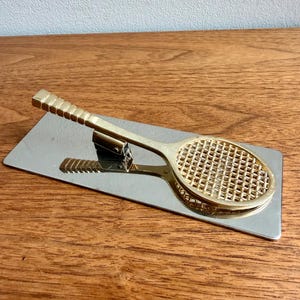 May include: A gold-colored tennis racket-shaped letter opener resting on a silver rectangular base. The handle of the letter opener has a textured grip. The base is on a wooden surface.