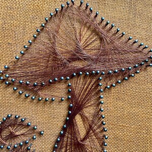 Pode incluir: Uma pe&ccedil;a de arte de cordas que representa um cogumelo com um fundo de juta marrom. O cogumelo &eacute; feito com fio de cobre e tachinhas azuis.
