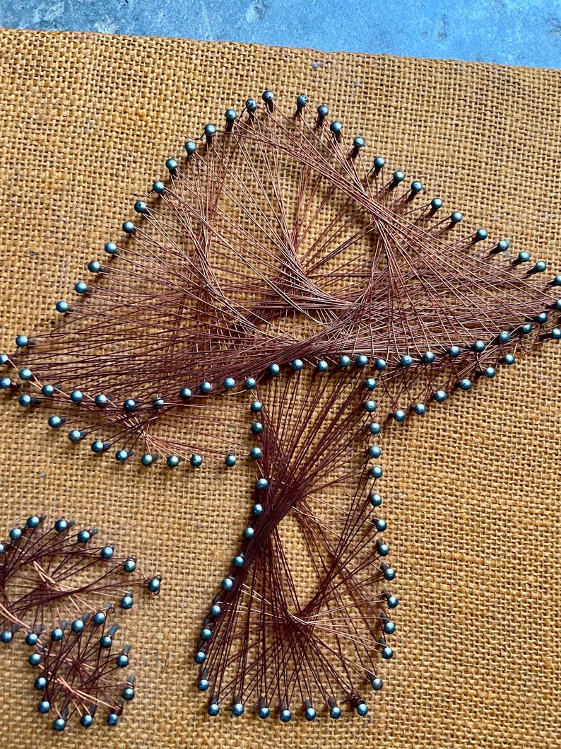 Pode incluir: Uma pe&ccedil;a de arte de cordas que representa um cogumelo com um fundo de juta marrom. O cogumelo &eacute; feito com fio de cobre e tachinhas azuis.