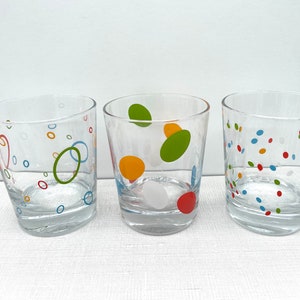 May include: Three clear drinking glasses with colorful dot and circle patterns. The glasses have a slightly angled design and are decorated with red, blue, green, and orange shapes. The glasses are suitable for water, juice, or other beverages.