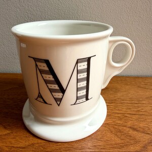 May include: A white ceramic mug with a large black letter "M" on the front. The letter is decorated with horizontal lines. The mug has a small handle and sits on a matching white saucer.