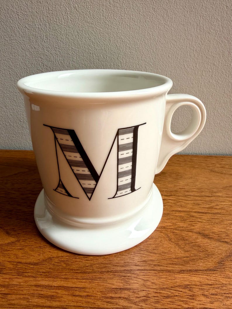May include: A white ceramic mug with a large black letter "M" on the front. The letter is decorated with horizontal lines. The mug has a small handle and sits on a matching white saucer.