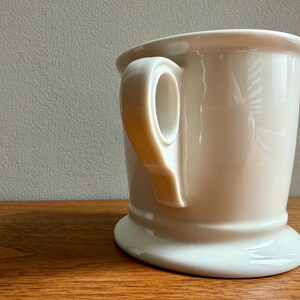May include: A cream-colored shaving mug with a rounded base and a large handle. The mug is made of ceramic and sits on a wooden surface. The background is a neutral gray.