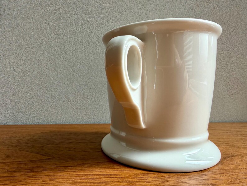 May include: A cream-colored shaving mug with a rounded base and a large handle. The mug is made of ceramic and sits on a wooden surface. The background is a neutral gray.