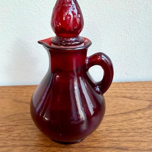 May include: A ruby red glass cruet with a teardrop-shaped stopper. The cruet has a small handle and a spout. The stopper has a textured surface. The cruet is sitting on a wooden surface.