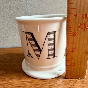 May include: White ceramic mug with a large black letter "M" on the front. The letter is decorated with a striped pattern. A wooden ruler is next to the mug. The mug is approximately 3 inches tall.