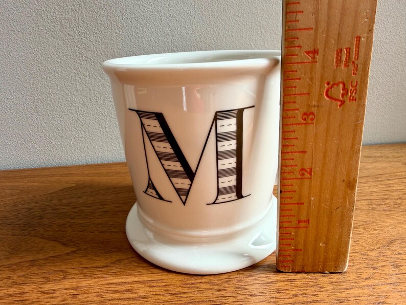 May include: White ceramic mug with a large black letter "M" on the front. The letter is decorated with a striped pattern. A wooden ruler is next to the mug. The mug is approximately 3 inches tall.