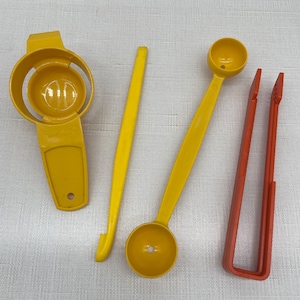 May include: A collection of yellow kitchen utensils, including an egg separator, a stirring tool, two measuring spoons, and a pair of orange tongs. The tools are made of plastic and are arranged on a white surface.