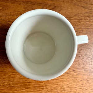 May include: A white ceramic mug with a handle, viewed from above. The mug is empty and sits on a wooden surface. The interior of the mug is smooth and clean, with a slightly rounded base.