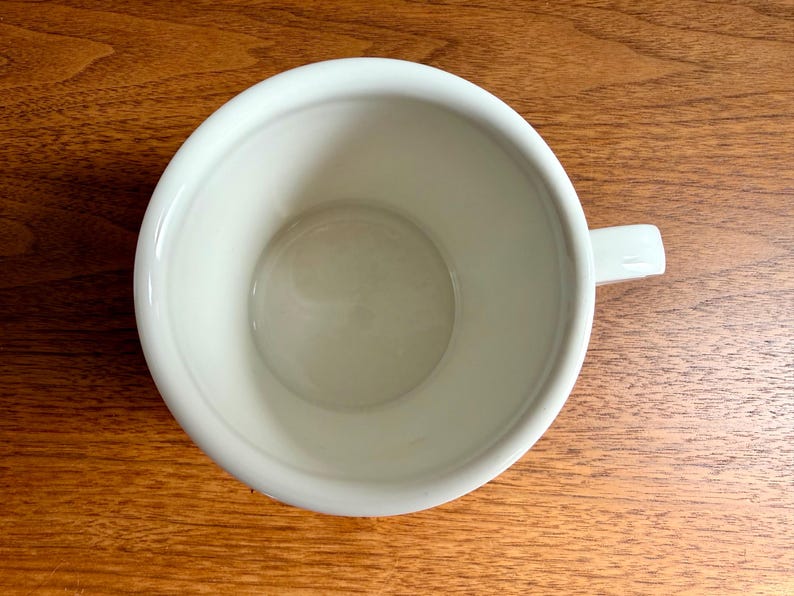 May include: A white ceramic mug with a handle, viewed from above. The mug is empty and sits on a wooden surface. The interior of the mug is smooth and clean, with a slightly rounded base.