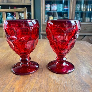 May include: Two ruby red goblets with a textured, geometric pattern. The goblets have a rounded base, a stem, and a flared bowl. They are displayed on a wooden surface, showcasing their rich color and design.