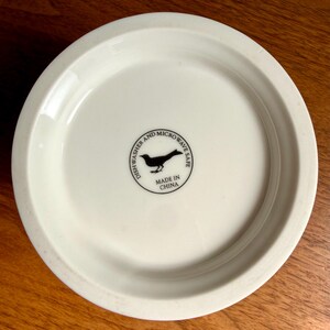 May include: Off-white ceramic plate with a slightly raised edge. The bottom of the plate features a black bird silhouette within a circle, along with the text "Dishwasher and Microwave Safe" and "Made in China".