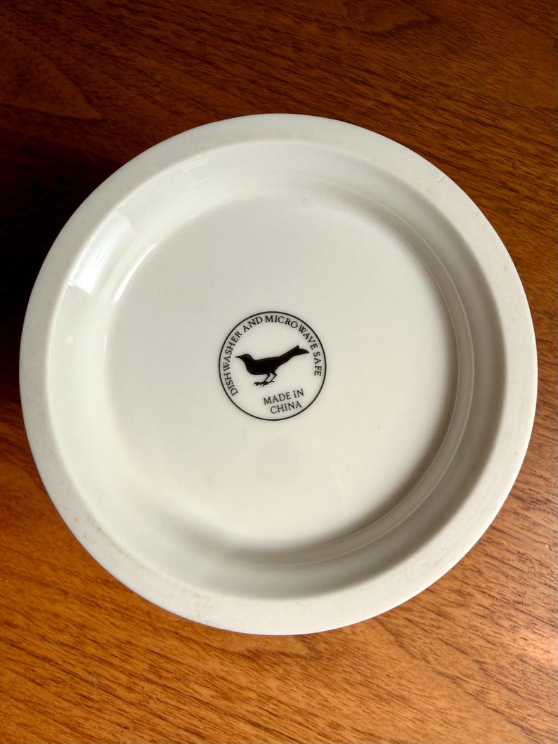 May include: Off-white ceramic plate with a slightly raised edge. The bottom of the plate features a black bird silhouette within a circle, along with the text "Dishwasher and Microwave Safe" and "Made in China".
