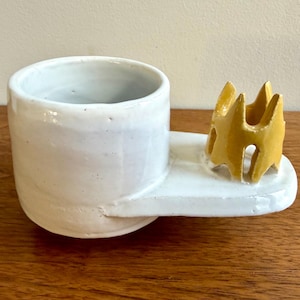 May include: A handmade ceramic piece with a white cup and a white rectangular base. A yellow, abstract, crown-like shape is attached to the base. The cup and base are glazed, and the piece sits on a wooden surface.