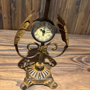 May include: A gold-toned metal table clock featuring a round face with black Roman numerals and hands. The clock is mounted on a decorative stand with leaf-shaped side accents, a square base with radial ridges, and a flat circular top platform.