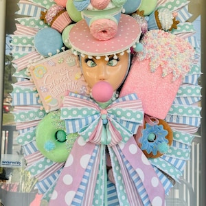 Op de afbeelding: Een grillige krans met een poppengezicht, versierd met pastelkleurige donuts, cupcakes en ijs. De krans bevat een grote strik en de tekst "Have a Sweet Christmas".