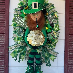 Puede incluir: Un duende verde sosteniendo una olla de monedas de oro, con un sombrero verde y zapatos verdes. El duende está rodeado de tréboles verdes y dorados y cintas. El texto "Happy St. Patrick's Day" y "Prone to Shenanigans & Malarkey" es visible en la corona.