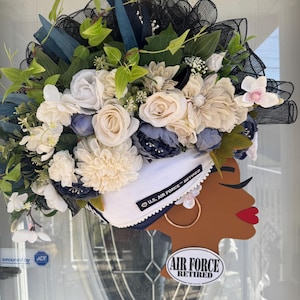 Peut inclure: Une couronne décorative avec une silhouette en bois de femme aux lèvres rouges et boucles d'oreilles dorées. La couronne est ornée de fleurs blanches, bleues et beiges, de verdure et de tulle noir. Le texte "U.S. AIR FORCE RETIRED" est visible.