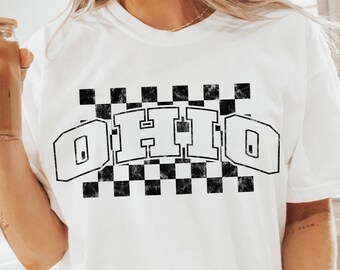 Op de afbeelding: Wit T-shirt met een zwart-wit geruit ontwerp en het woord "OHIO" in vetgedrukte letters. Het shirt wordt gedragen door een persoon die op een wit oppervlak zit.