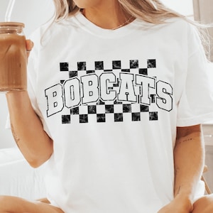 May include: White t-shirt with a black and white checkered design and the text "BOBCATS" in a retro font.