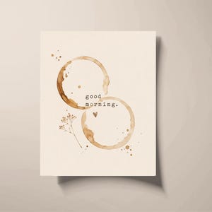 May include: A rectangular card featuring two overlapping brown coffee mug stains on a cream background. The text "good morning." is printed in black typewriter font between the rings, accompanied by a small hand-drawn heart and a sprig of baby's breath.