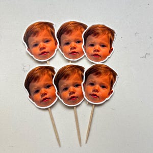 May include: Six cupcake toppers featuring a baby's face with light brown hair and a white border. The toppers are attached to wooden sticks, arranged in two rows of three on a white surface.