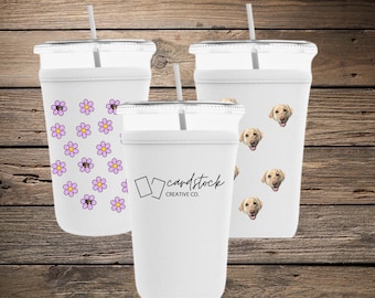 May include: Three white plastic tumblers with lids and straws. One tumbler has a pattern of pink and purple flowers. Another has images of a dog's face. The third tumbler has the text "cardstock CREATIVE CO."
