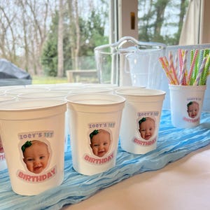 May include: White plastic cups with a personalised sticker featuring a baby's photo and the text "Zoey's 1st Birthday".