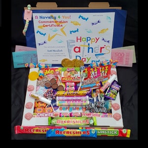 Könnte beinhalten: Eine blaue und weiße Karte mit dem Text "Happy Father's Day" und ein Zertifikat für "Scott Mcculloch", umgeben von einer Vielzahl bunter Süßigkeiten, Lutscher und Schokoladenriegel in einer weißen Schachtel.