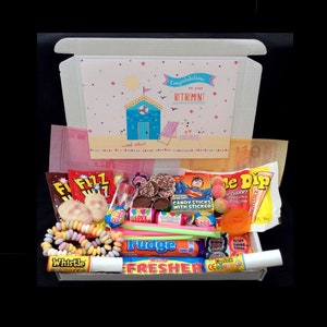 Pode incluir: Uma caixa de presente branca cheia de uma variedade de doces coloridos, incluindo moedas de chocolate, palitos de doces, pirulitos e gomas. A caixa tem um cartão no topo que diz "Parabéns pela sua aposentadoria...e relaxe!" com uma cena de praia e uma cabana de praia azul.