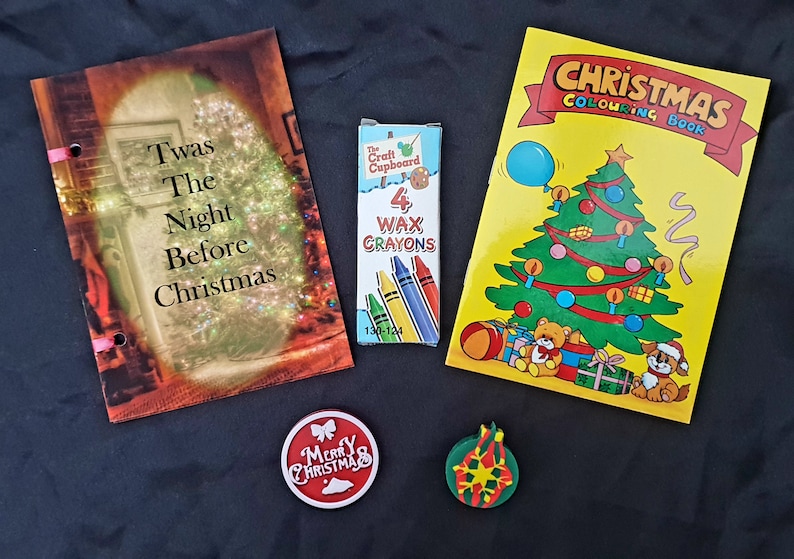 May include: A Christmas-themed gift set with a book titled "Twas The Night Before Christmas", a coloring book with a Christmas tree and a teddy bear, a box of four wax crayons, a red ornament with the text "Merry Christmas", and a green ornament with a snowflake design.
