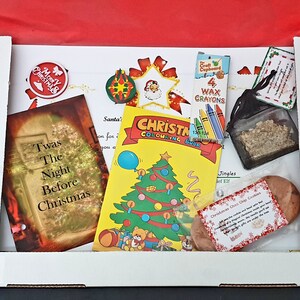 May include: A white box filled with Christmas-themed items, including a coloring book, a small book titled "Twas The Night Before Christmas", a bag of glitter, a small bag of cookies, and a set of wax crayons. The box is decorated with a red ribbon.