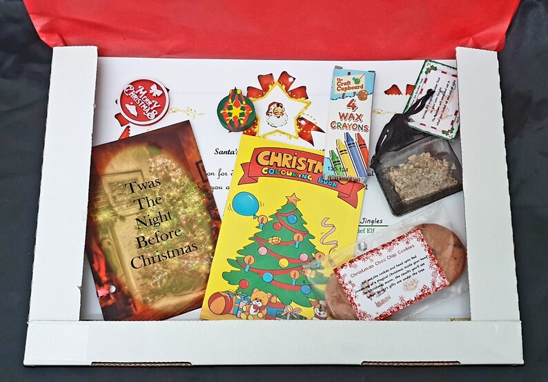 May include: A white box filled with Christmas-themed items, including a coloring book, a small book titled "Twas The Night Before Christmas", a bag of glitter, a small bag of cookies, and a set of wax crayons. The box is decorated with a red ribbon.
