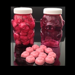 May include: Two pink plastic jars shaped like pigs, filled with pink candy hearts. The jars are sitting on a table with a scattering of pink candy hearts in front of them.