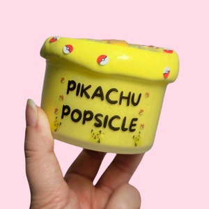 Op de afbeelding: Gele slijm in een container met de tekst "Pikachu Popsicle" erop. De container is versierd met rode en gele Pok&eacute; Balls.