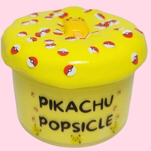 Op de afbeelding: Gele slijm met een Pikachu-ijsje erin en rode en witte Pokeball-sprinkels erop. De slijm zit in een doorzichtige container met de tekst "Pikachu Popsicle" op de voorkant.