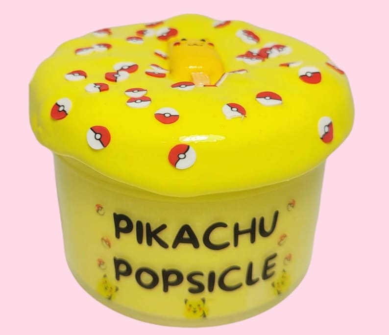 Op de afbeelding: Gele slijm met een Pikachu-ijsje erin en rode en witte Pokeball-sprinkels erop. De slijm zit in een doorzichtige container met de tekst "Pikachu Popsicle" op de voorkant.
