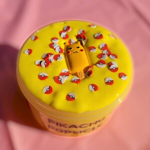 Op de afbeelding: Gele slime met rode en witte Pok&eacute;bal-sprinkels en een Pikachu-ijsje-bedel. De slime zit in een doorzichtige container met de tekst "Pikachu Popsicle" op het etiket.