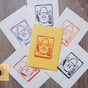 May include: A collection of linocut prints featuring a cartoon character wearing a cowboy hat and holding a can of soda. The prints are in various colors, including red, blue, orange, and black, on white and yellow paper.