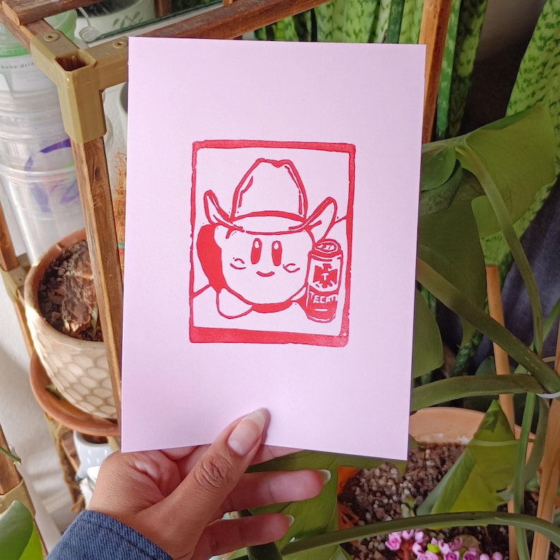 May include: A pink print featuring a red cartoon character wearing a cowboy hat and holding a can of Tecate beer. The character is inside a red square. The print is on pink paper.