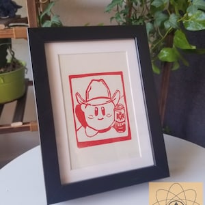 May include: Red and white framed print featuring a cartoon character wearing a cowboy hat and holding a can of Tecate beer. The print is set against a white mat and framed in a black frame. The artwork is displayed on a white surface.