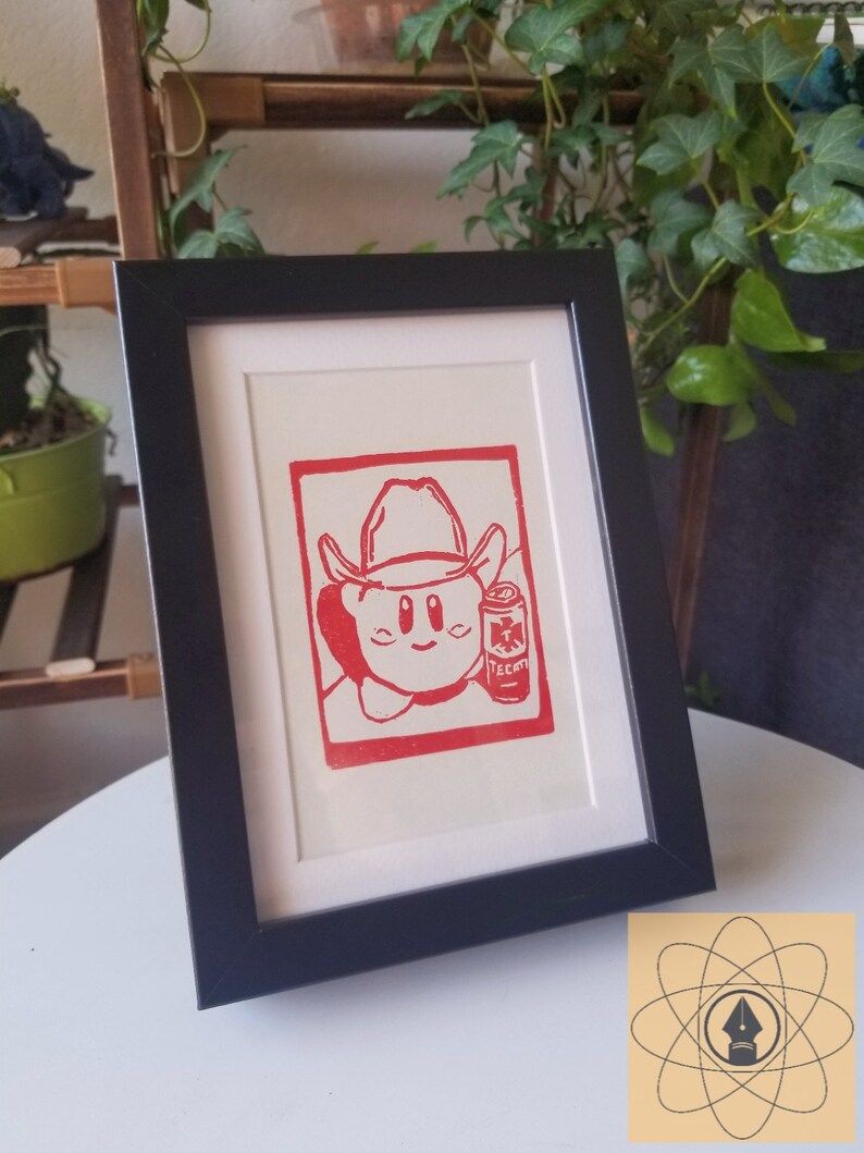 May include: Red and white framed print featuring a cartoon character wearing a cowboy hat and holding a can of Tecate beer. The print is set against a white mat and framed in a black frame. The artwork is displayed on a white surface.